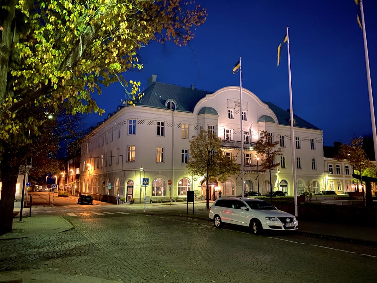 En gata framför en stor byggnad med gulvita fasader och upplysta fönster i skymningen. Till vänster syns ett träd med gulgröna löv. En mörk bil står parkerad på gatan i bakgrunden och en vit kombi är parkerad i förgrunden.