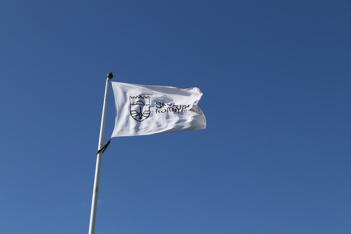En vit flagga med ett svart tryck föreställande ett vapen omgivet av texten "Skurups kommun" vajar från en vit flaggstång mot en klarblå himmel.