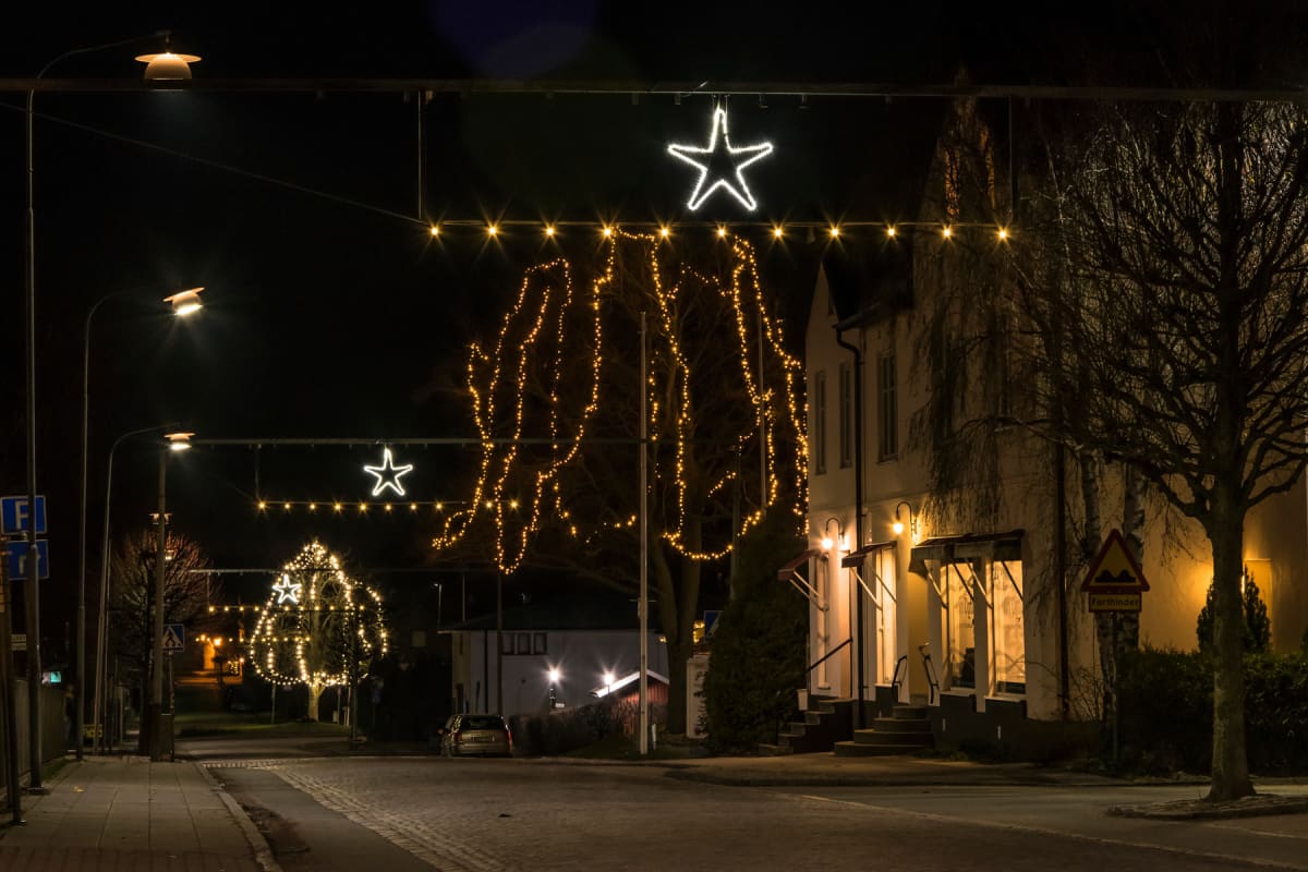 En gata kvällstid med julbelysning i form av stjärnor och ljusslingor som hänger över vägen. Träd längs gatan är dekorerade med ljusslingor. Längs ena sidan av gatan finns en upplyst byggnad med trappor och entréer. Vid trottoaren syns gatlyktor och