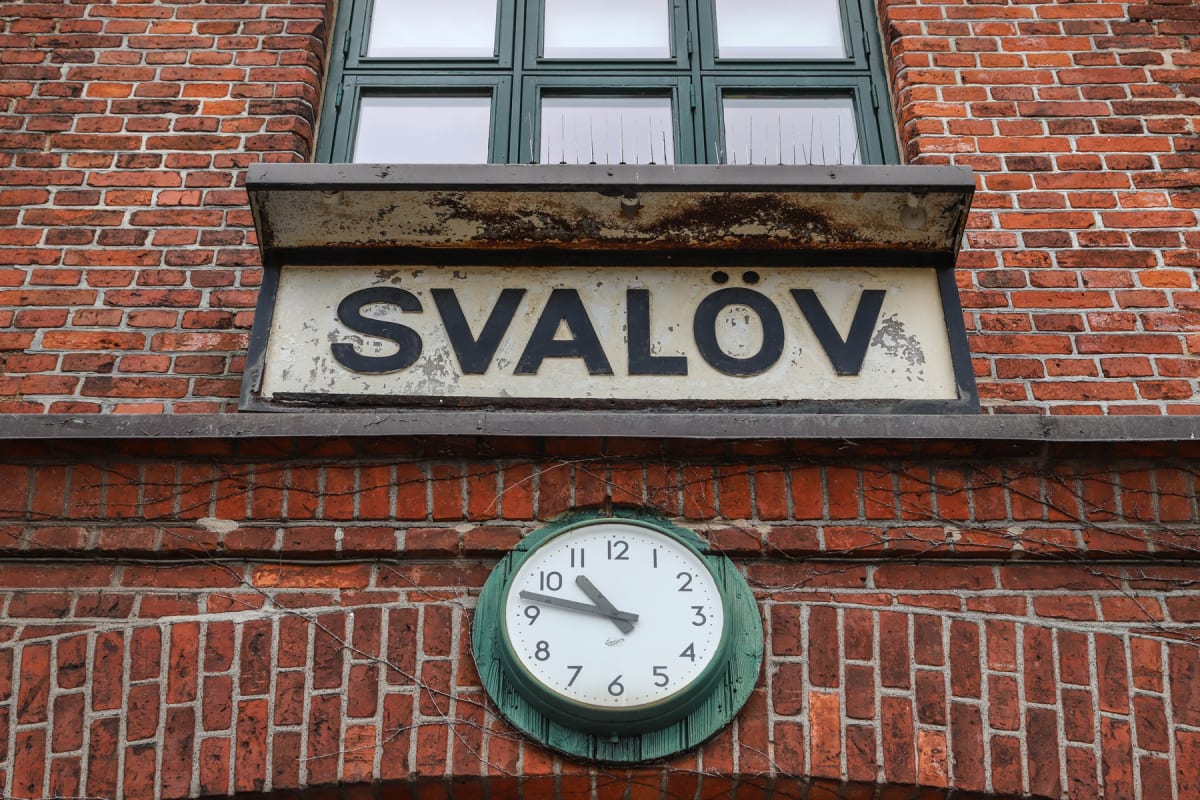 En fasad av rött tegel med ett stort fönster med mörkgröna spröjsar överst. Under fönstret sitter en skylt med texten "SVALÖV" i svarta bokstäver på en vit bakgrund. Längre ner på tegelfasaden sitter en rund grön klocka med vita siffror och svarta vi
