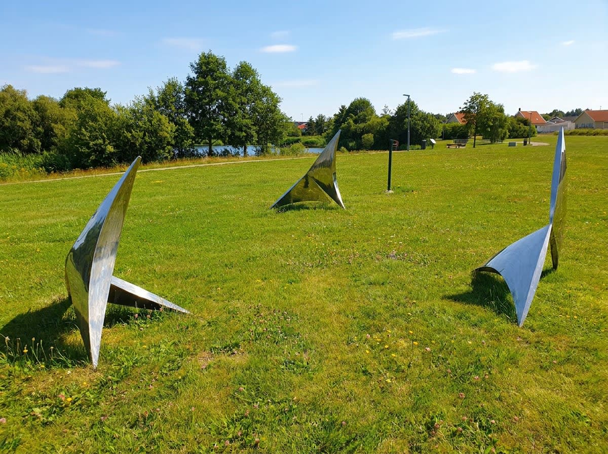 En utomhusbild som visar tre abstrakta, reflekterande metallskulpturer placerade i en grön gräsyta. I bakgrunden syns träd, buskar, en vattenyta och byggnader under en blå himmel med några moln.