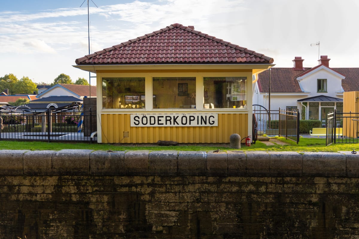 En gul byggnad med rött tak och vita fönsterkarmar mot en blå himmel med moln. En skylt med texten "SÖDERKÖPING" sitter på byggnadens fasad. Framsidan av byggnaden vetter mot en vattenyta som är kantad av en stenvägg. I bakgrunden syns fler byggnader