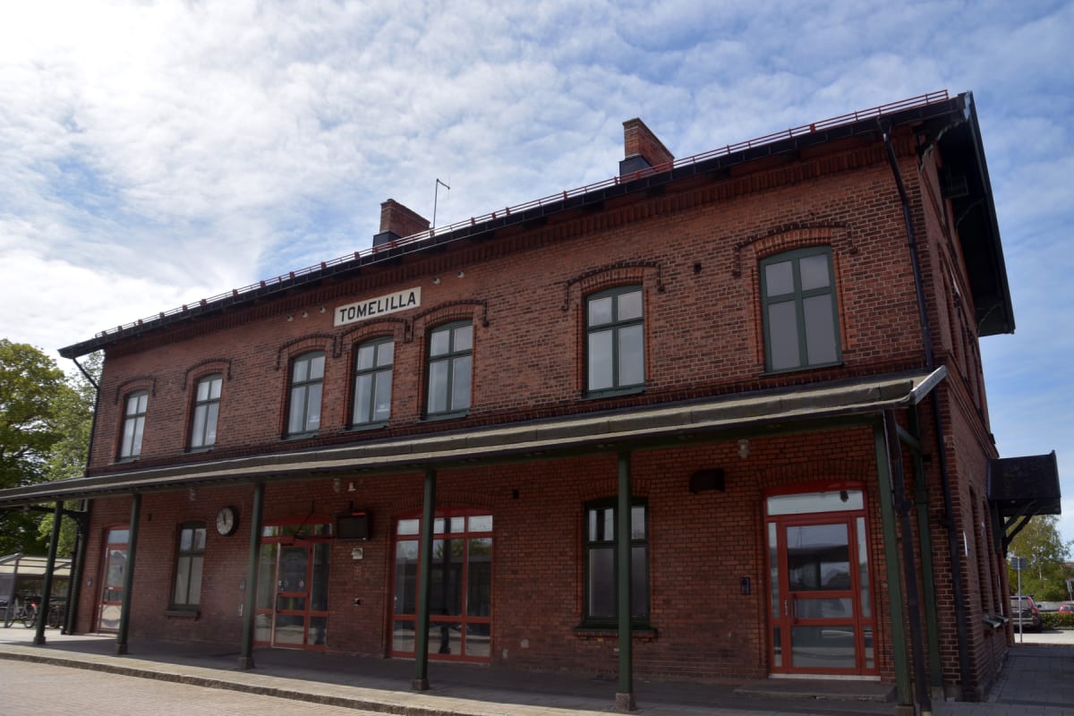 En stor, röd tegelbyggnad med många fönster och en plattform under ett utskjutande tak. Byggnaden har vita skyltar med texten "TOMELILLA" på fasaden. Himlen är blå med vissa moln och träd syns i bakgrunden.