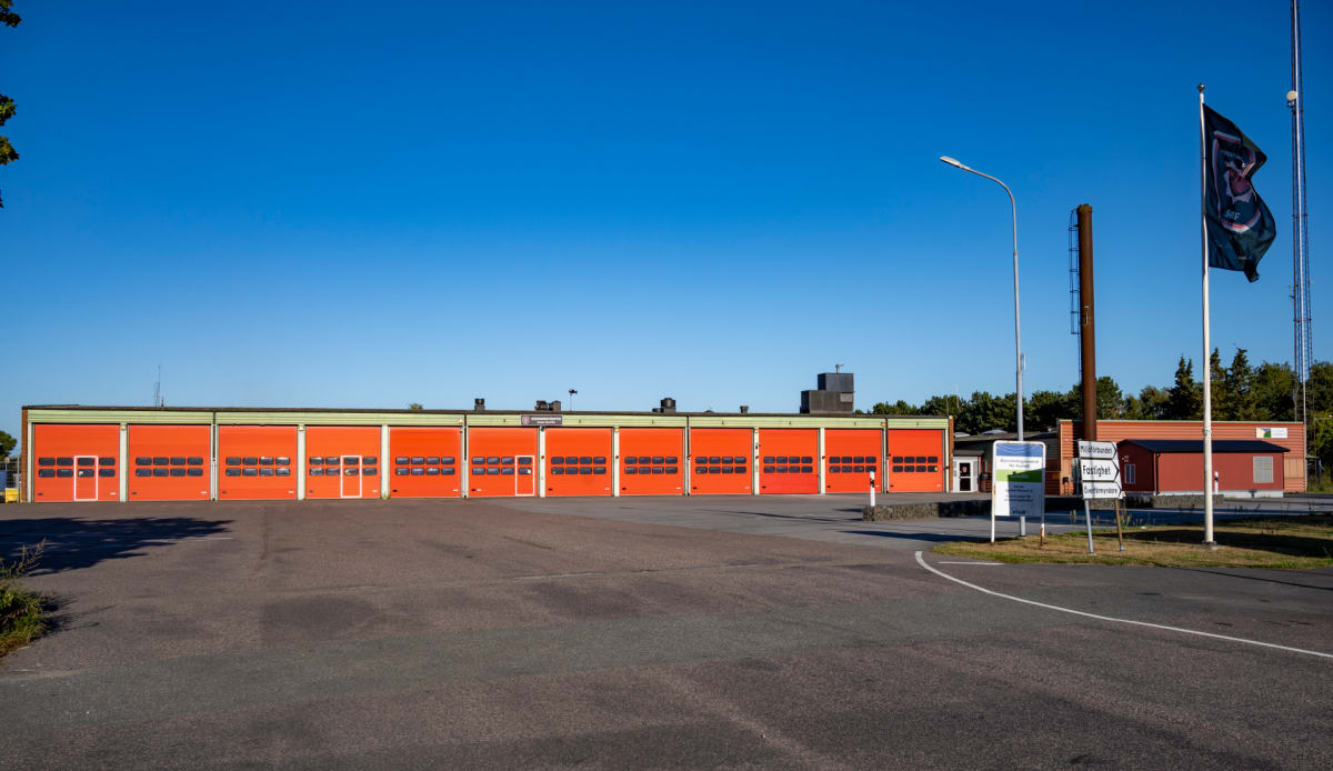 En byggnad med orangea portar under en klarblå himmel. Framför byggnaden finns asfalterad mark och till höger en flaggstång med en mörk flagga, en ljusstolpe och ett brunt torn. Några skyltar står vid infarten och träd syns i bakgrunden.