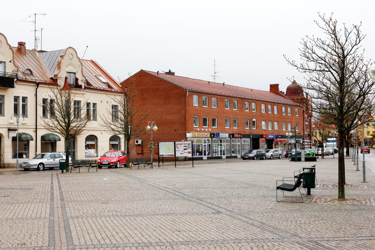 En ganska bred bild av ett torg i en stad med en kullerstensbelagd yta. Byggnader med tegeltak och fasader i gult och tegel omger torget. Flera bilar är parkerade längs byggnaderna och bare träd står utspridda. Himlen är grå och molnig.