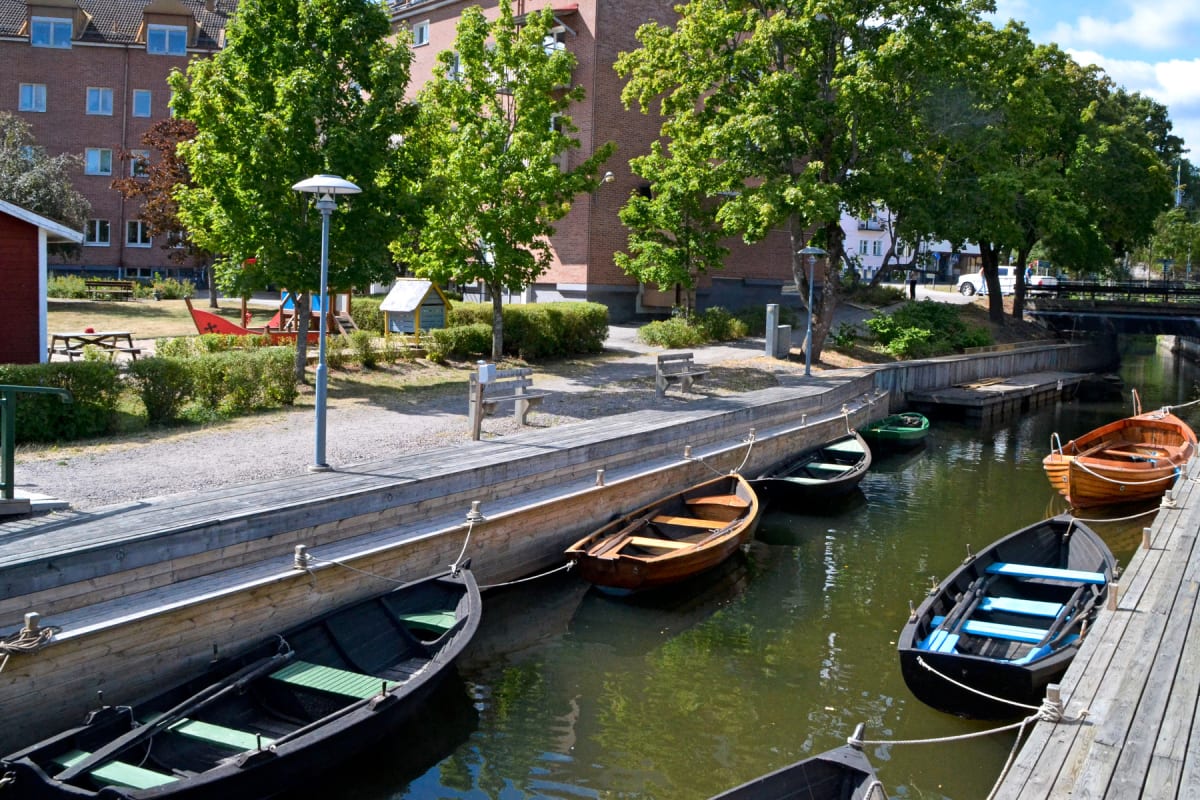 En bild som visar en kanal eller å med flera roddbåtar förtöjda längs en träbrygga och ett trädäck. I bakgrunden syns bostadshus i tegel, gröna träd och buskar samt en lekplats.