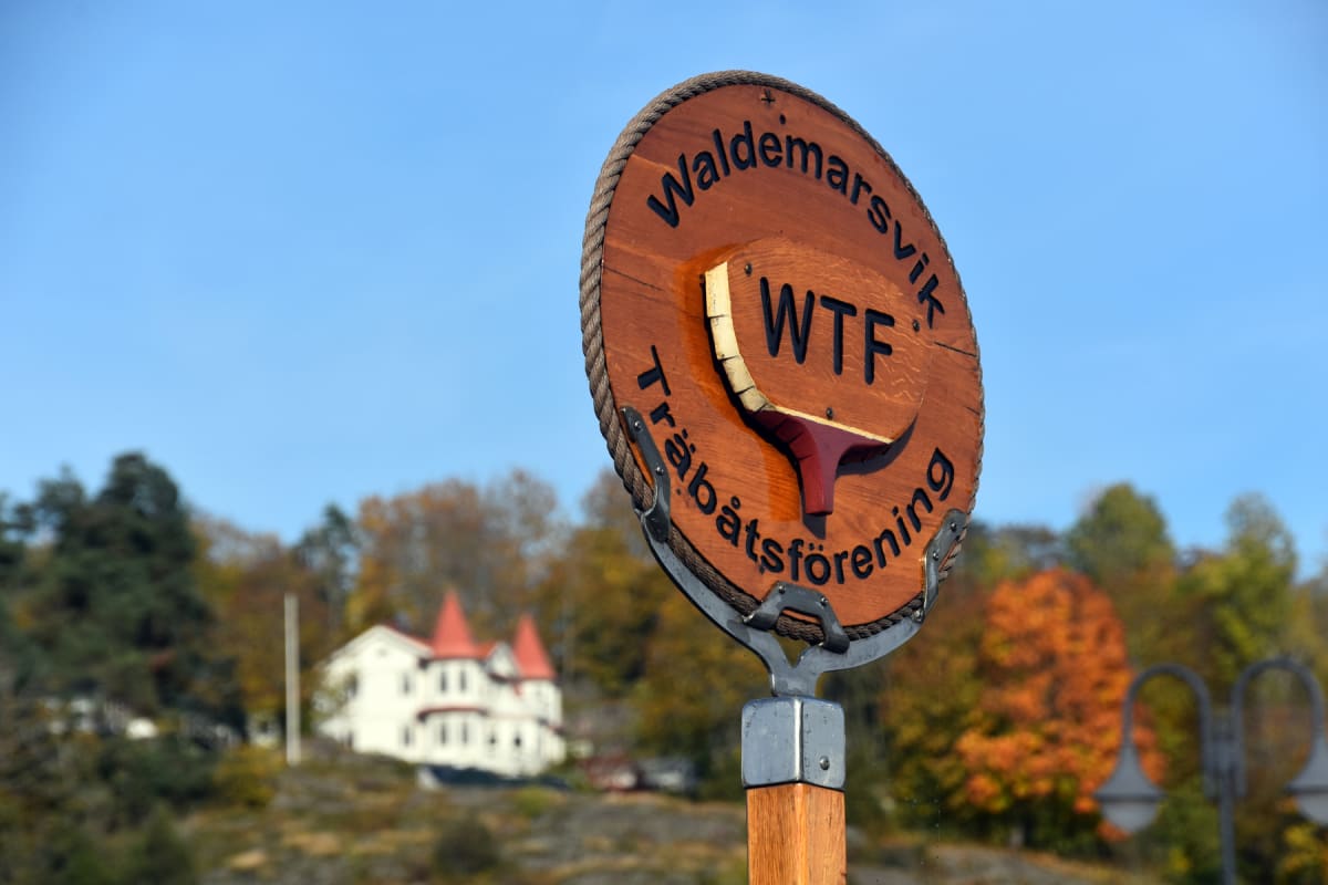 En närbild av en rund träskylt med texten ”Waldemarsvik” och ”Träbåtsförening” som inramar en symbol med bokstäverna ”WTF”. Skylten är monterad på en trästolpe. I bakgrunden syns skiftande träd i höstfärger, en vit byggnad med röda takdetaljer, och a