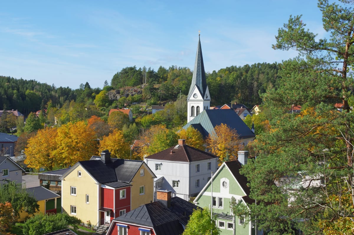 En översiktsbild av en stadsmiljö med en kyrka i centrum, omgiven av bostadshus och träd i höstfärger. De varierande trädens lövverk och en klarblå himmel bidrar till bilden. En tall är synlig i förgrunden till höger.