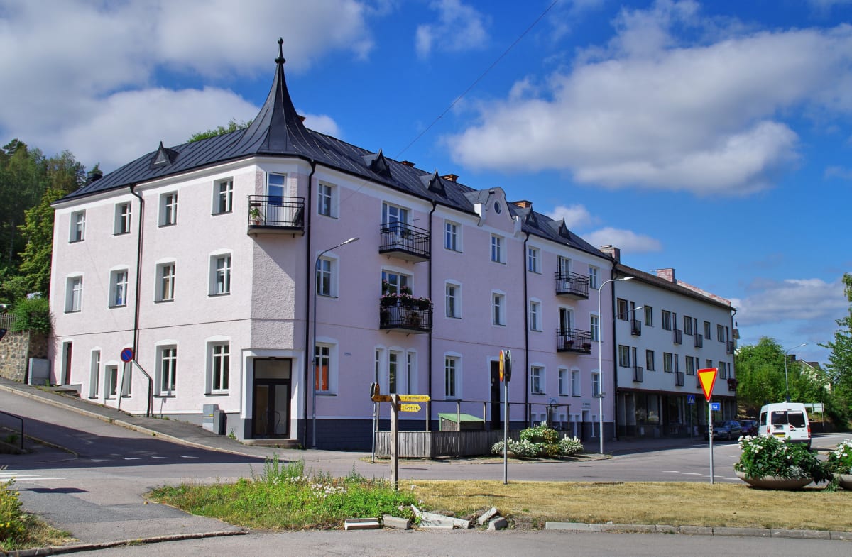 En ljusrosa byggnad med svart tak och balkonger står i förgrunden, omgiven av asfalt och gräs. I bakgrunden syns en annan byggnad, träd, samt en blå himmel med vita moln. Gul-svarta vägskyltar och lyktstolpar är synliga.