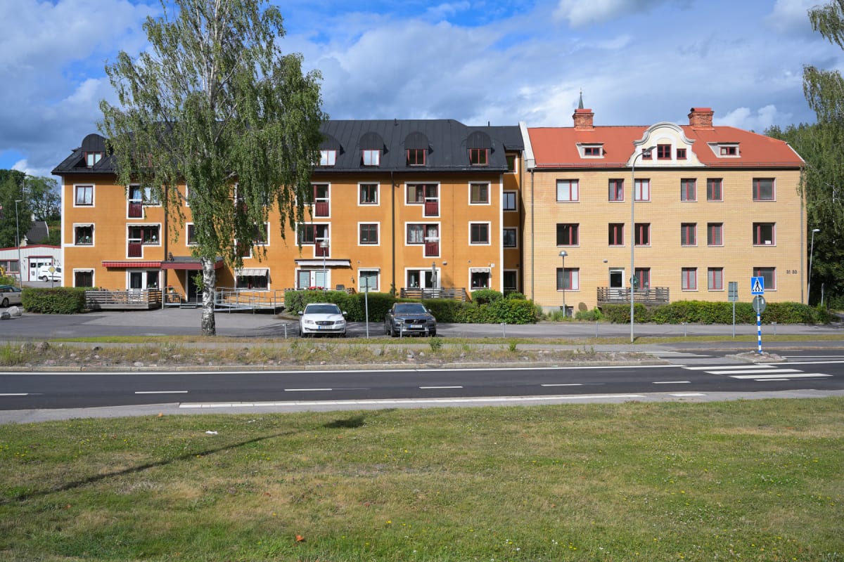 En bild tagen utomhus under dagtid som visar en gräsyta, en asfaltsväg med vita markeringar och i bakgrunden två flerfamiljshus i olika färger, det ena gult och det andra tegelfärgat. Flera bilar är parkerade framför husen.