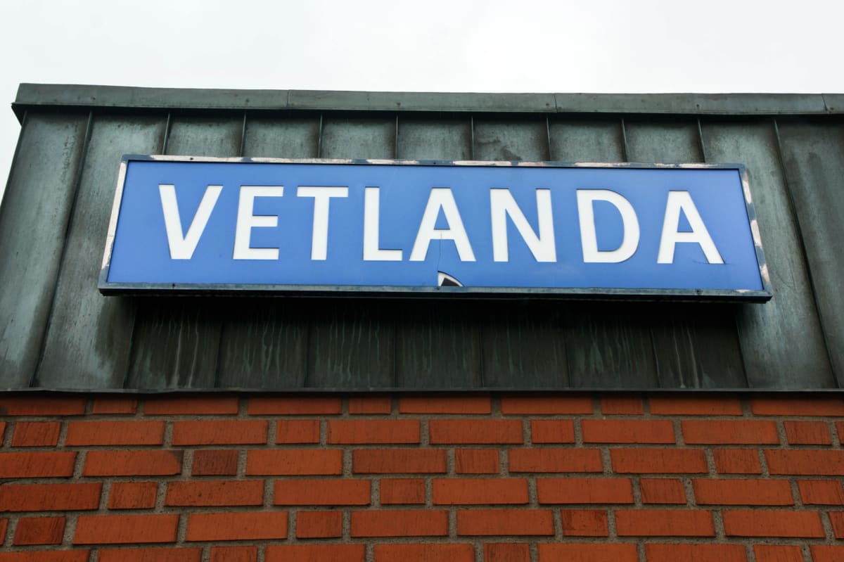 En blå skylt med vita bokstäver där texten "VETLANDA" står är monterad under en mörk takfot. Skylten hänger ovanför en tegelvägg. Himmel syns i bakgrunden.