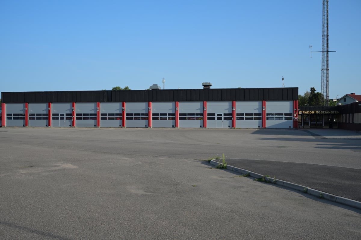 En bild som visar framsidan av en byggnad med flera garageportar i två våningar. Utanför byggnaden finns ett asfalterat område och i bakgrunden syns skog och en mast. På höger sida av byggnaden finns en skylt med texten RÄDDNINGSTJÄNSTEN.