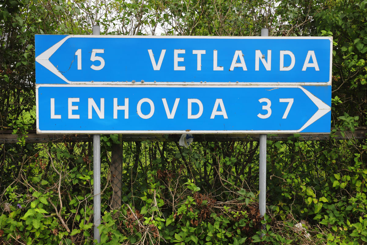 En blå vägskylt med vit text som visar riktning till Vetlanda 15 och Lenhovda 37. Skylten står mot en bakgrund av grönska och ett länkstängsel, med en ljus himmel ovanför. Texten är tydlig och skylten är monterad på en metallstolpe.