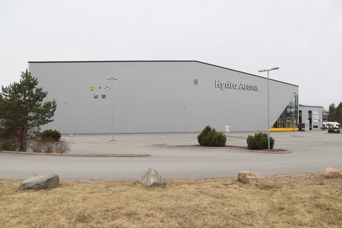 En stor, grå byggnad märkt "Hydro Arena" med ett parkeringsområde framför. Framför byggnaden finns ett torkat gräsområde med några stenar och buskar. Himlen är grå och mulen. På byggnadens vänstra sida syns flera små, runda emblem.