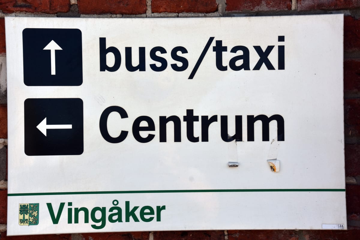 En vägvisningsskylt fäst på en tegelvägg. Skylten är vit med svart text som anger "buss/taxi" med en uppåtpil och "Centrum" med en vänsterpil. Längst ner finns Vingåkers kommunlogga och namnet "Vingåker". Skylten visar spår av slitage.