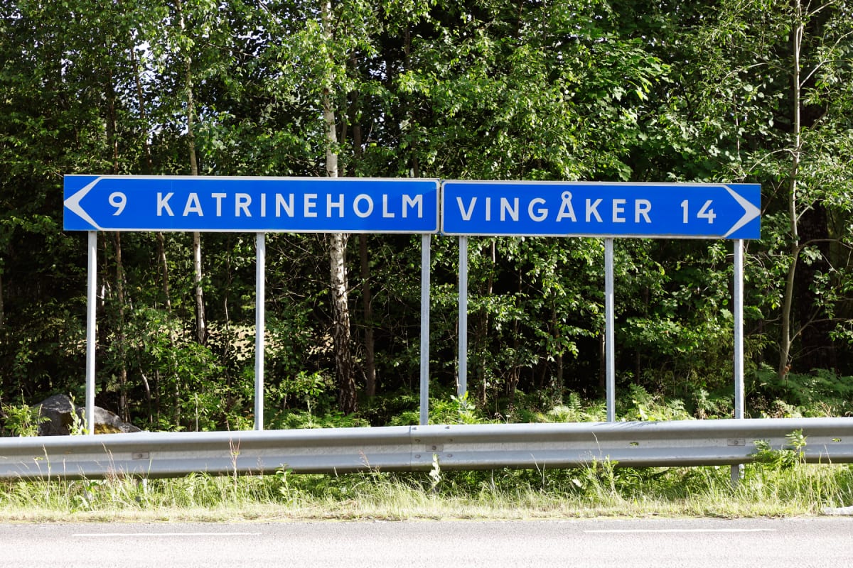 En blå vägvisningsskylt hänger på sex stolpar och visar "9 Katrineholm" med en vänsterpil och "Vingåker 14" med en högerpil. Skylten står vid en väg med ett grått vägräcke framför en bakgrund av gröna träd och buskar.