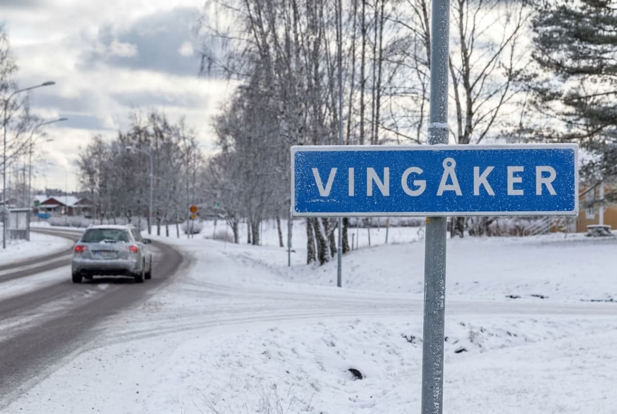 En blå vägskylt med texten "VINGÅKER" står vid sidan av en snötäckt väg. En silverfärgad bil kör iväg på vägen som svänger till vänster. Snö täcker marken och grenarna på träden, och himlen är molnig.