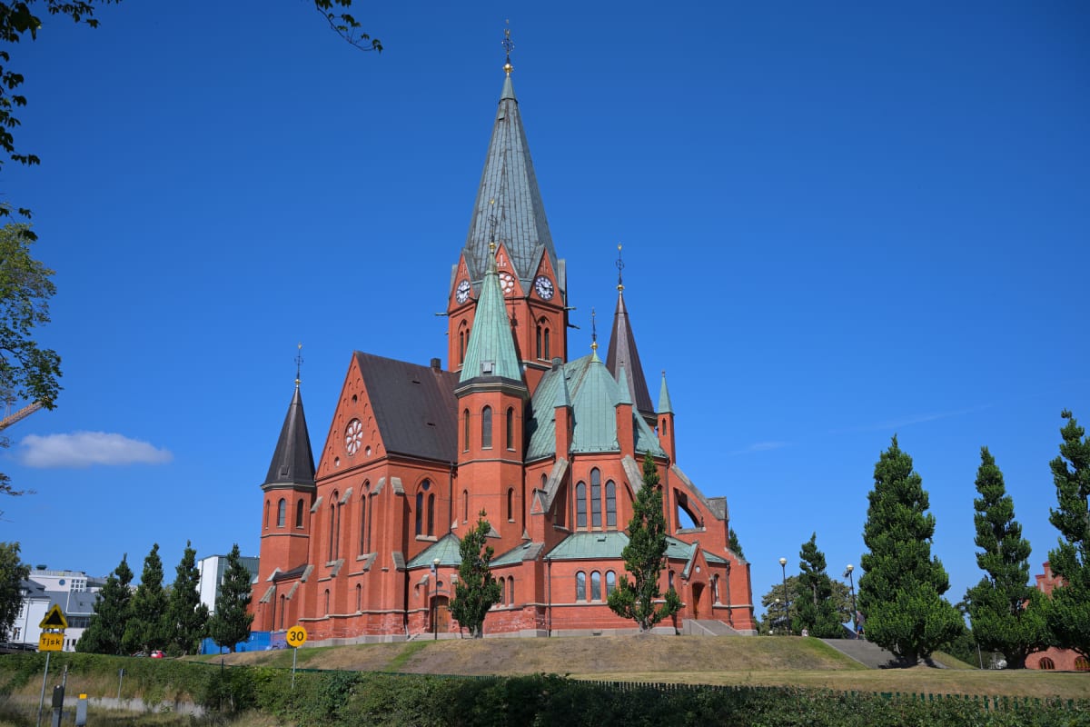 En stor, röd tegelkyrka med flera spetsiga torn och gröna koppartak under en klarblå himmel. Runt kyrkan finns grönska i form av buskar, gräs och träd. I bakgrunden syns delar av byggnader och en gul varningsskylt med silhuetten av en person som sitt