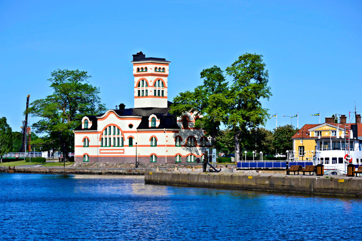 En bild tagen i dagsljus som visar en vit byggnad med orange och gröna detaljer och en hög tornliknande del. Byggnaden står vid en vattenkant. Till höger syns en gul byggnad och en vit båt. Gröna träd och en blå himmel syns i bakgrunden.
