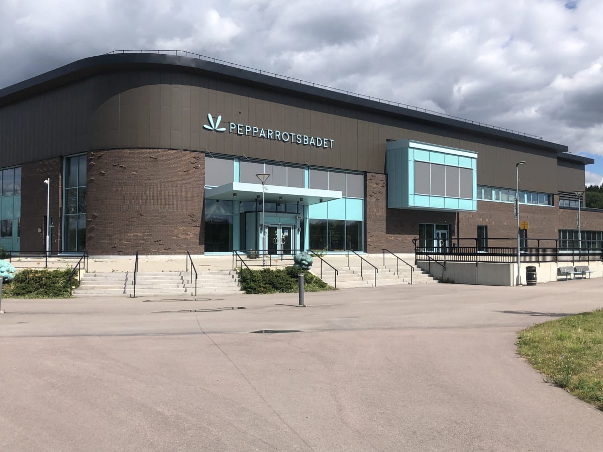 En modern byggnad med en entré med stora glaspartier och en skylt där det står "PEPPARROTSBADET". Byggnaden har fasad i tegel och mörkbruna skivor, samt turkosfärgade detaljer. Framför entrén finns en trappa samt stenlagd yta med gräs.