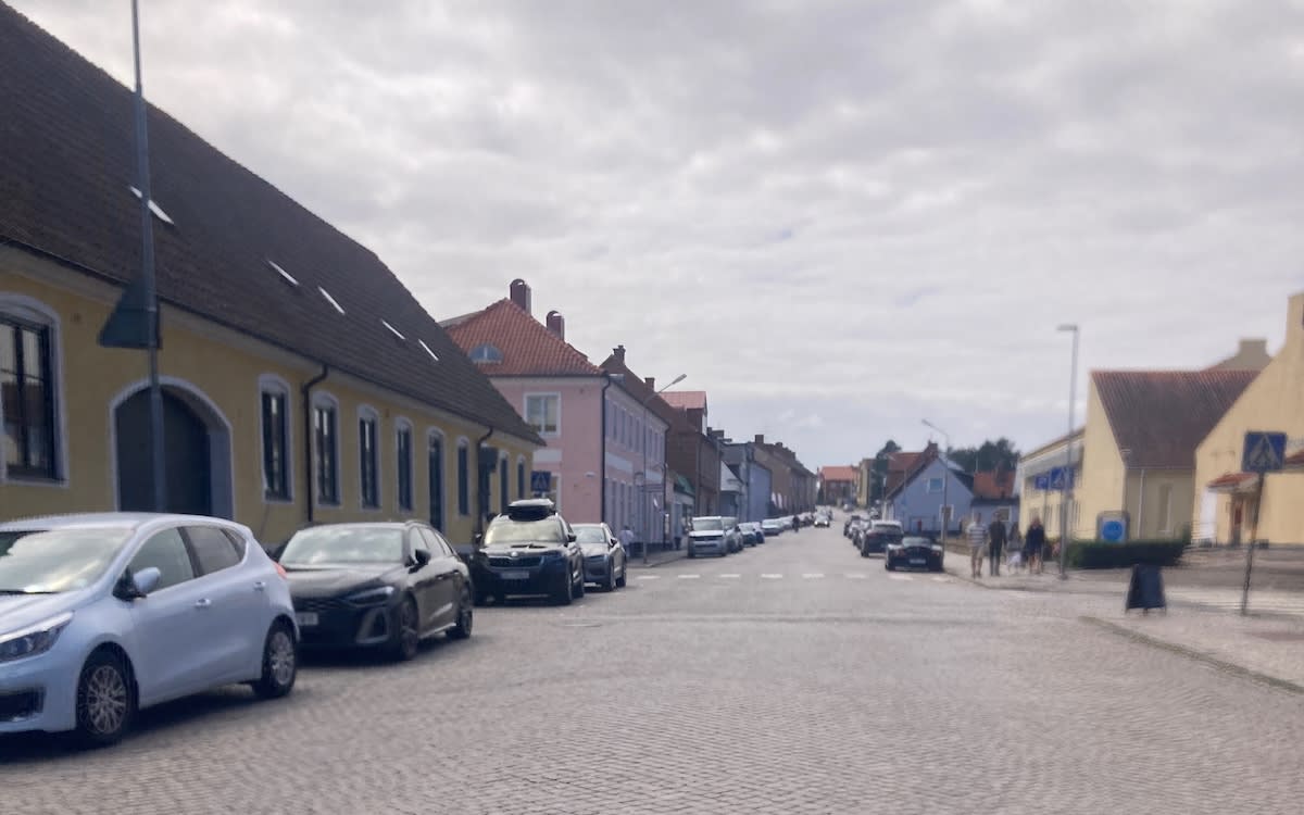 En gata i en stad med kullersten och byggnader längs sidorna. Bilar står parkerade längs gatan och några människor går på trottoaren. Himlen är molnig och det finns gatljus uppsatta.
