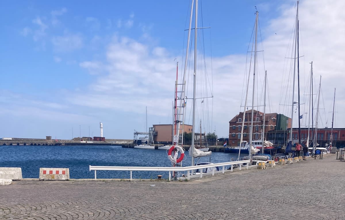 En bild som visar en småbåtshamn med flera segelbåtar förtöjda vid en kaj. I bakgrunden syns havsvatten, en vågbrytare med en fyr, samt byggnader. Himlen är blå med några moln. En livboj är fäst vid en stolpe i förgrunden.