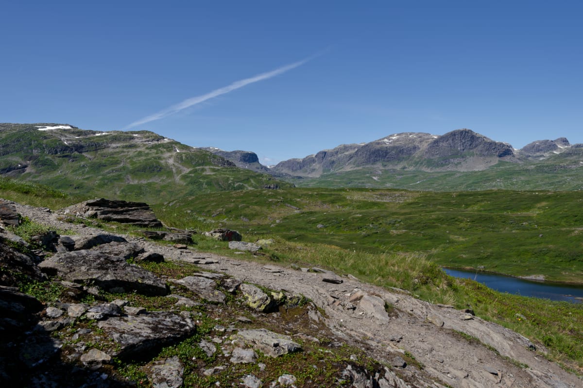 En vidvinkelbild tagen utomhus under dagtid, som visar ett kuperat, bergigt landskap med grön vegetation och en ljusblå himmel. Ett spår av kondens från ett flygplan syns på himlen. I förgrunden syns stenar, mark och vegetation.