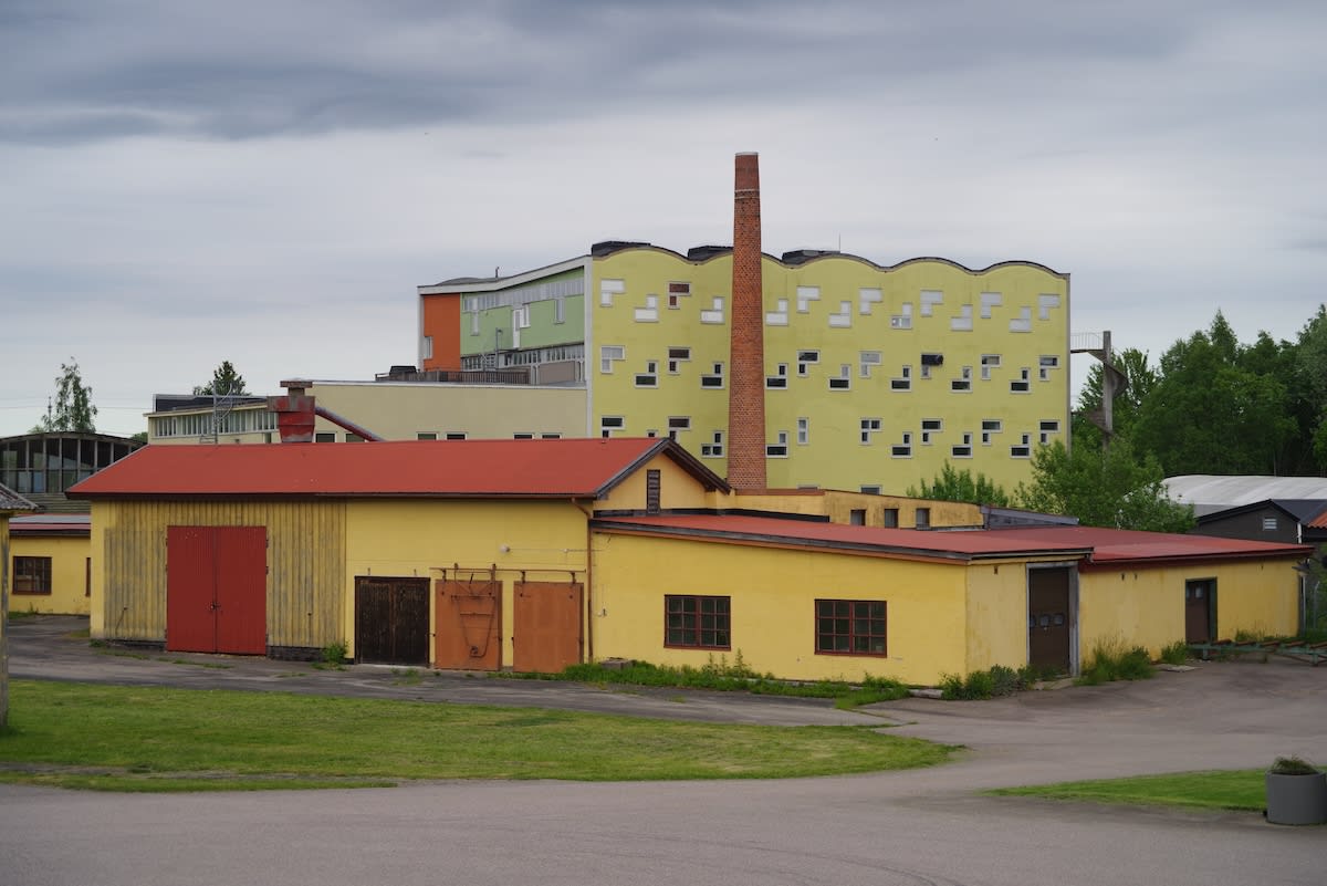 En bild som visar en samling med byggnader i olika färger och former, under en mulen himmel. I förgrunden syns gulmålade byggnader med röda tak och portar. I bakgrunden reser sig större byggnader, bland annat en gul byggnad med en skorsten. Grönskand