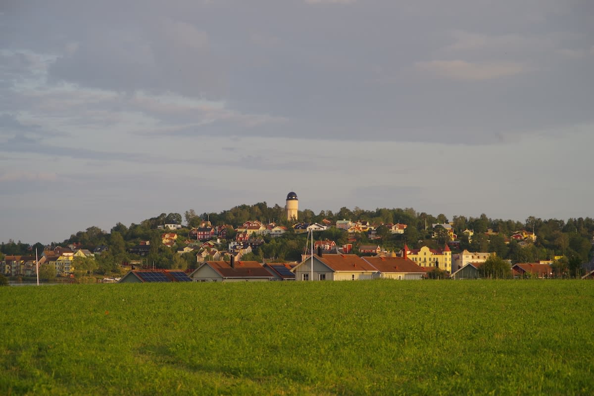 En bild som visar ett grönskande fält i förgrunden, följt av fler hustak och byggnader på en kulle. På toppen av kullen syns en beige byggnad med ett mörkt, kupolformat tak.