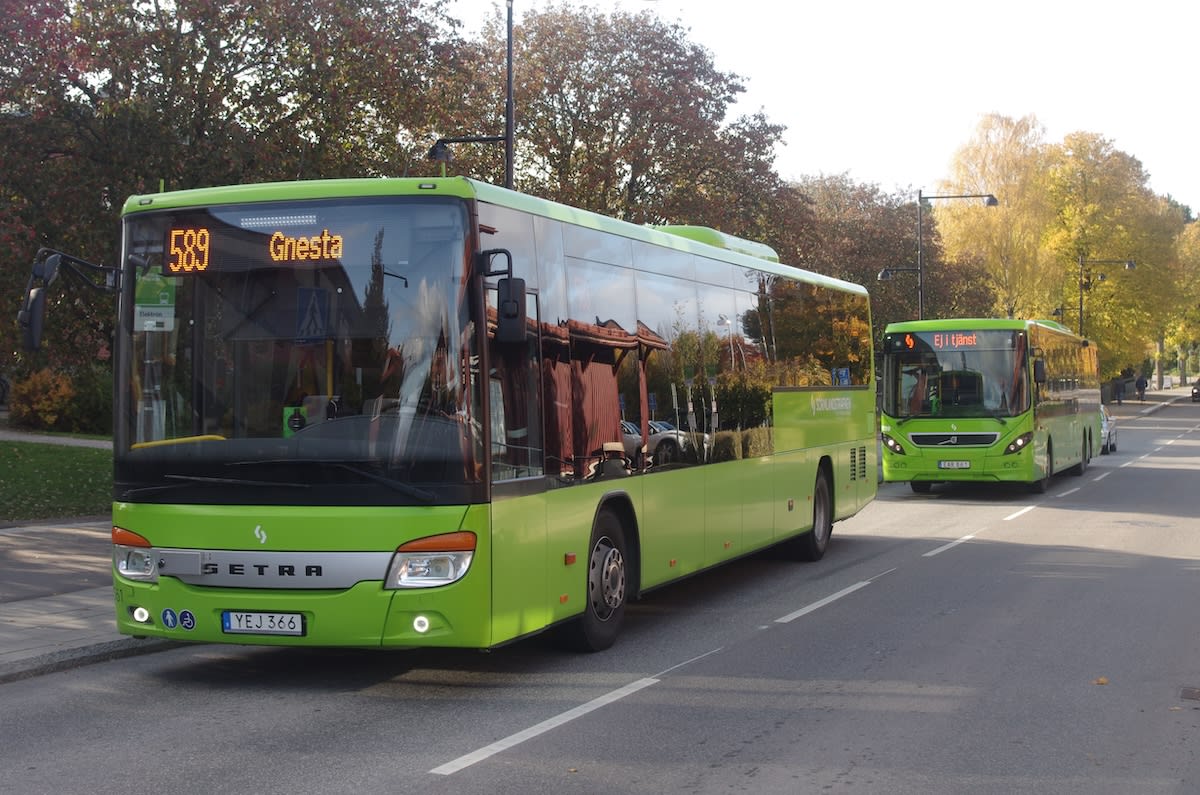 En bild som visar en grön buss med texten "589 Gnesta" på den främre destinationstavlan, parkerad vid sidan av en väg. I bakgrunden syns en annan grön buss som kör på vägen. Träd med höstlöv omger området.
