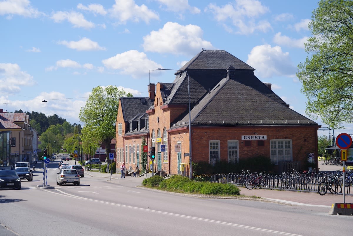 En gata med biltrafik under dagtid. Vid sidan av gatan finns en stor byggnad i tegel med texten "GNESTA" på fasaden. Ett flertal parkerade cyklar står utanför byggnaden. Några personer syns på trottoaren. Grönska och träd omger byggnaderna. Himmelen