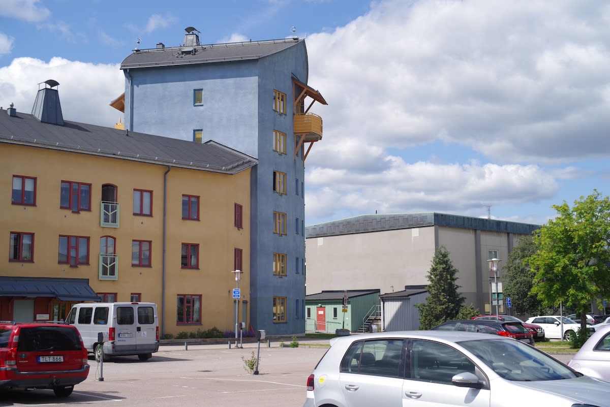 En bild som visar en parkeringsplats med flera parkerade bilar och en van. I bakgrunden syns gul- och blåfärgade flerbostadshus med röda fönsterkarmar samt en större, ljusare byggnad. Grönska från träd och buskar är synlig.