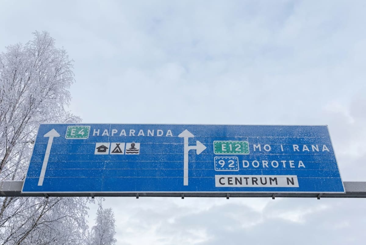 En blå vägskylt med vita pilar och text som indikerar riktningar mot Haparanda, Mo i Rana och Dorotea. Hela skylten och de närliggande träden är täckta av ett lager frost, vilket indikerar kallt väder. Bakgrunden består av en ljusgrå himmel med moln.