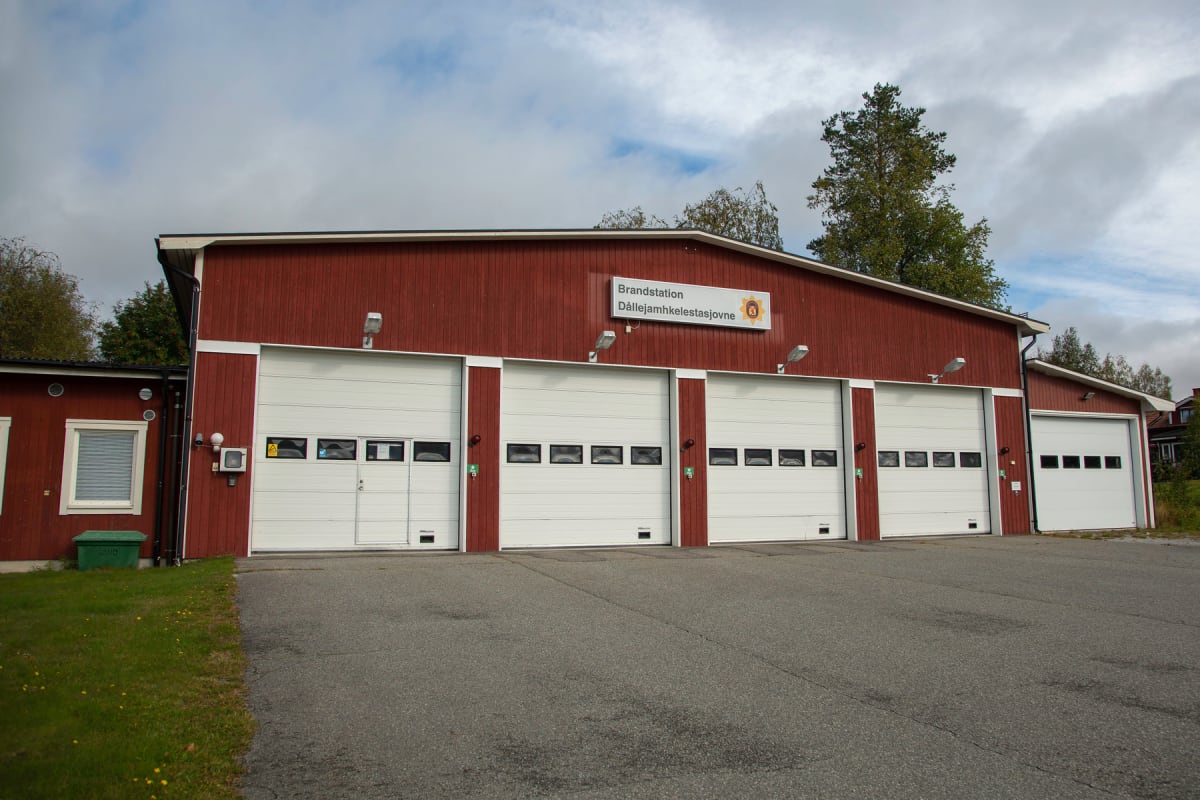 En röd brandstation med vita portar under en molnig himmel. Byggnaden har sex vita garageportar, varav fem är breda och försedda med fönster. En skylt ovanför centrala portarna indikerar "Brandstation Dállejamhkelestastajovne". Asfalt framför byggnad