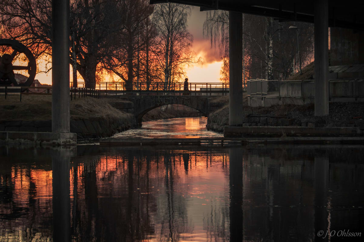 En bild som visar en person som går över en bro vid solnedgången, med trädsiluetter mot en orange himmel. Broar och vegetation reflekteras i vattnet. Ett vattenhjul syns till vänster. Pelare sträcker sig ner i vattnet i förgrunden.