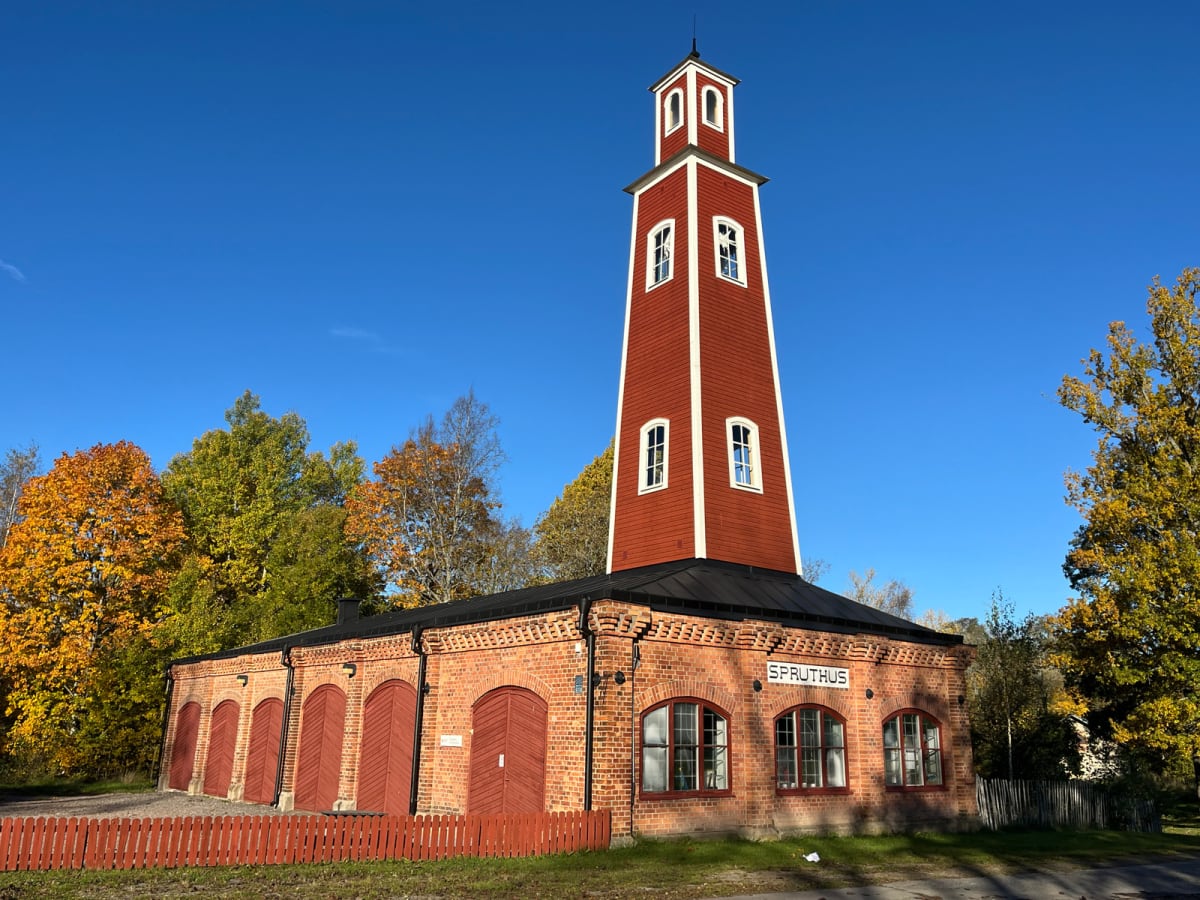 En röd tegelbyggnad med en hög, spetsig, röd trä-tornbyggnad på taket, med vita fönsterkarmar mot en klarblå himmel. Runt byggnaden växer träd med löv i gula och gröna höstfärger, och framför tegelbyggnaden syns ett rött trästaket.