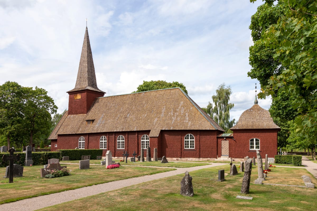 En röd träkyrka med en spetsig spira står mitt i bilden, omgiven av en kyrkogård med gräs och grusgångar, samt träd i bakgrunden. Klocktornet och del av huvudbyggnaden syns på vänster sida, ett mindre kapell på höger sida.