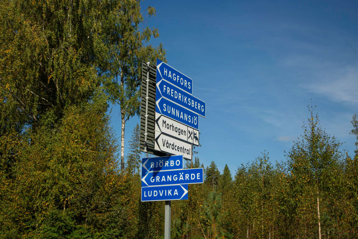 En vertikal bild visar en skylt med flera riktningspilar och ortsnamn. Skylten står vid en vägkant med lövträd i höstfärger och barrträd i bakgrunden. En klarblå himmel syns ovanför träden.