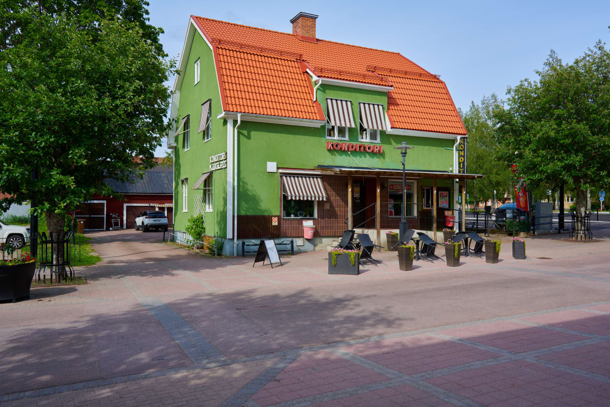 En ljusgrön byggnad med orange tak står i centrum av bilden. På byggnadens fasad syns texten "KONDITORI" i rött samt "KONDITORI & MEJERI" i vitt. Framför byggnaden finns ett uterum med bord och stolar. Till vänster i bilden står ett grönt träd och i