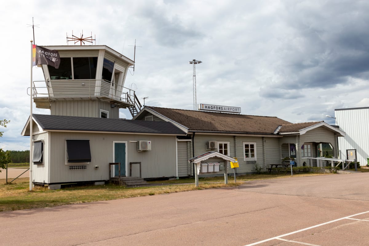 En bild av en flygplatsbyggnad med ett kontrolltorn under en molnig himmel. Byggnaden är ljust grå med vita knutar och mörka tak. En skylt med "HAGFORS AIRPORT" syns på taket. En flagga med texten "HAGFORS AIRPORT" flyger vid kontrolltornet. En väg o
