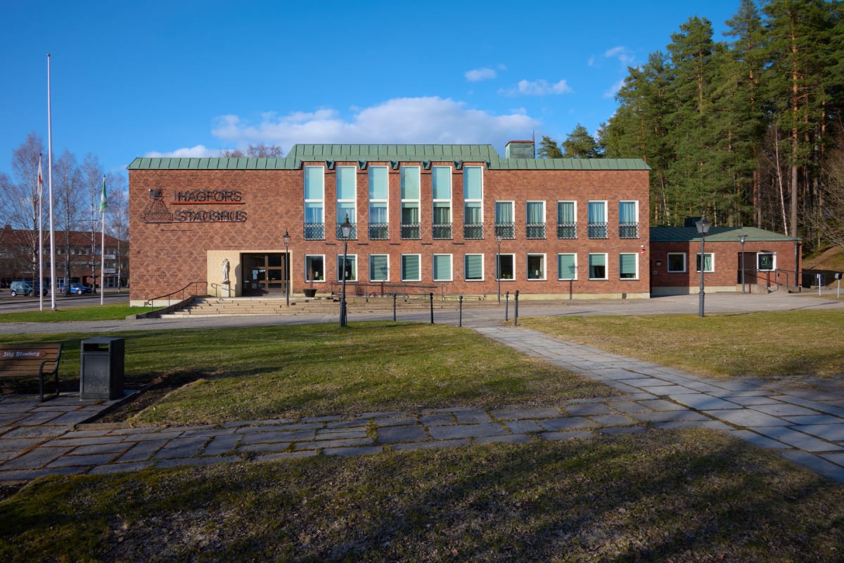 En stor, röd tegelbyggnad med texten "HAGFORS STADSHUS" är placerad i bildens mitt. Byggnaden har många fönster och en grön taklist. Framför byggnaden finns en putsad stengång som leder till ingången, samt gräsytor och lampstolpar. I bakgrunden syns