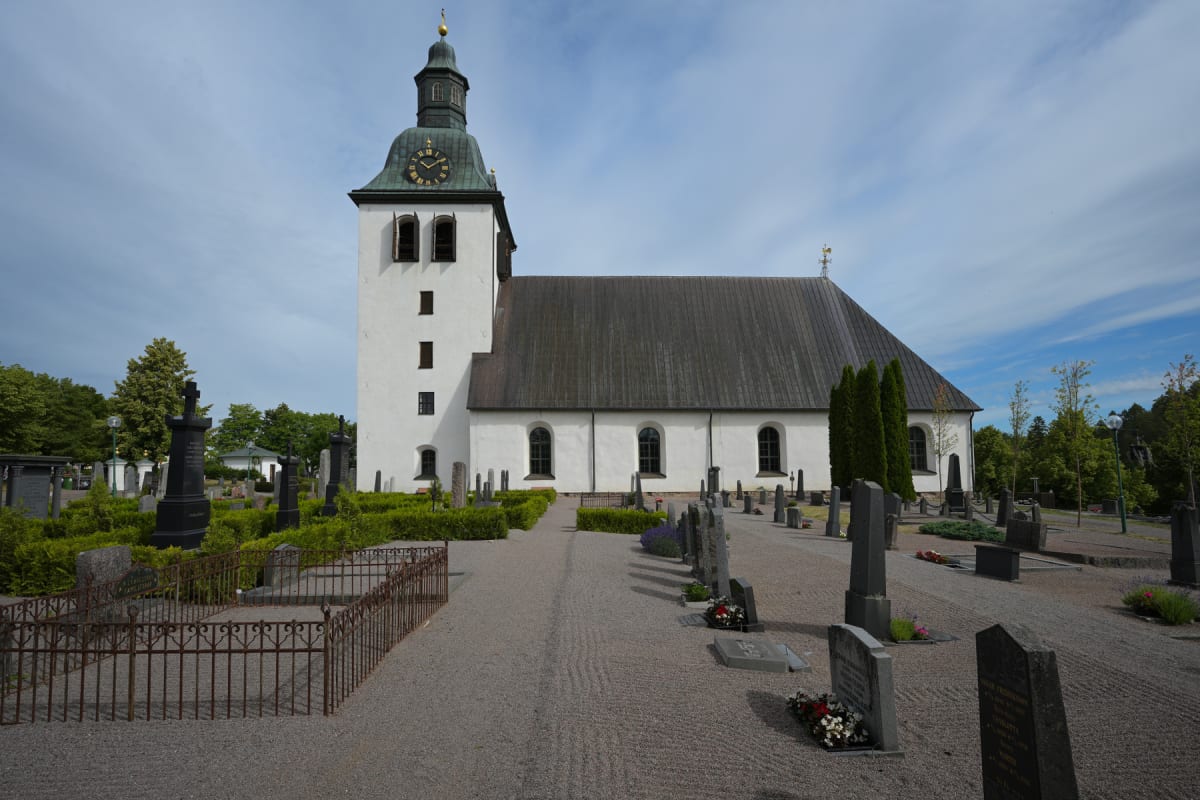 En vit kyrka med ett grått tak och ett högt klocktorn med ett ur. Kyrkan är omgiven av en kyrkogård med gravstenar och grusgångar, samt gröna buskar och träd under en delvis molnig himmel.