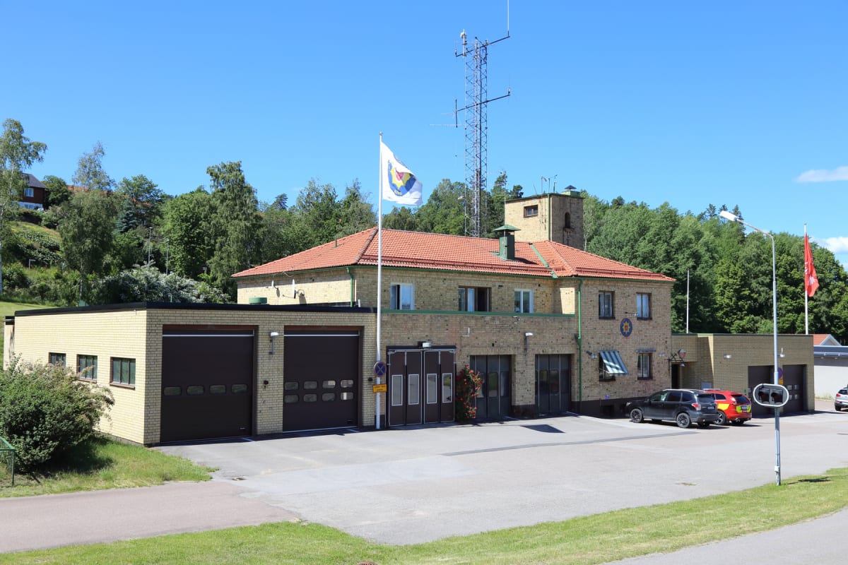 En gul tegelbyggnad med rött tak, som ser ut som en brandstation, under en klarblå himmel. Flera garageportar syns, en mast med antenner står bakom byggnaden och två flaggor vajar. Två bilar står parkerade utanför och trädgrönska omger byggnaden.
