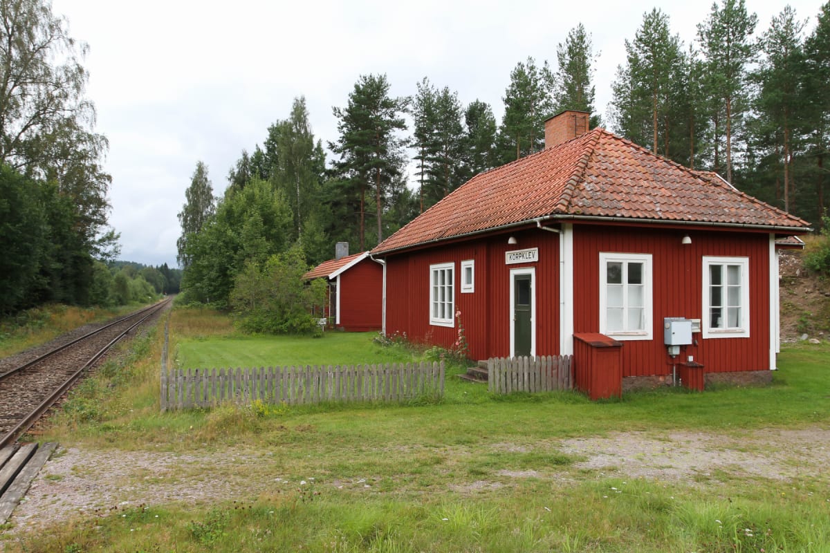 En röd byggnad med vita fönster och rött tak står intill en järnväg. Bredvid den röda byggnaden finns en mindre röd byggnad, också med rött tak. En trägärdsgård avgränsar gräsmattan. Skog syns i bakgrunden och himlen är molnig.