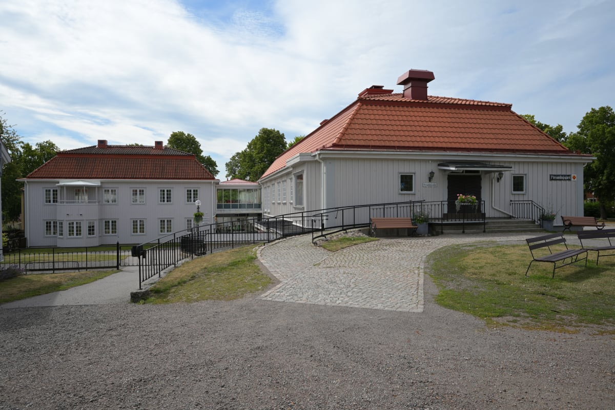 En bild visar två ljusa byggnader med röda tak under en molnig himmel. En grusväg i förgrunden leder till en gångväg av kullersten bland gräsytor. Flera bänkar står utplacerade i gräset och vid byggnadernas ingångar. Mellan byggnaderna finns en glask