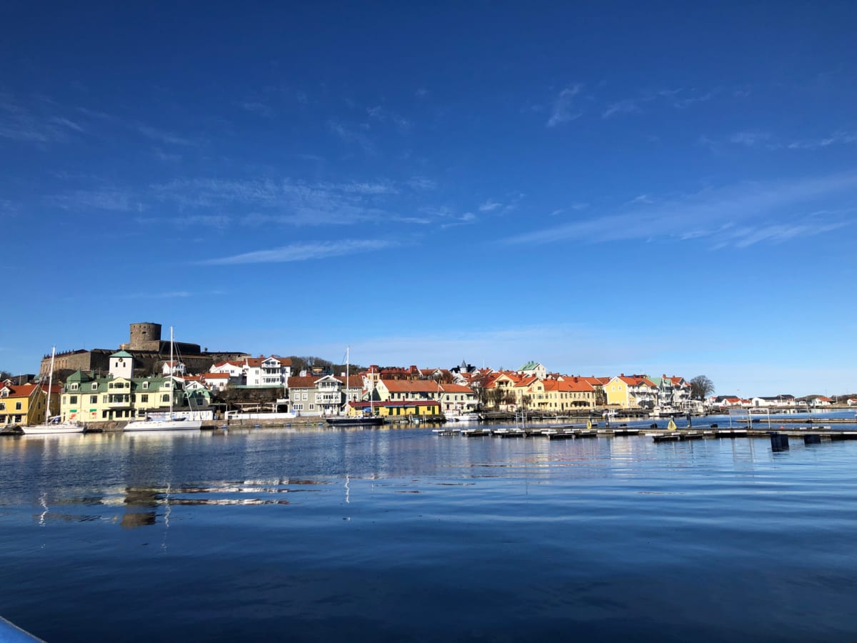 Bild tagen från vattenytan, visar en ljusblå himmel med tunna vita moln över en bebyggelse. Flera färgglada hus med orangea och röda tak syns längs strandlinjen, och i bakgrunden finns en stenfästning. Båtar ligger förtöjda vid bryggor på vattnet.