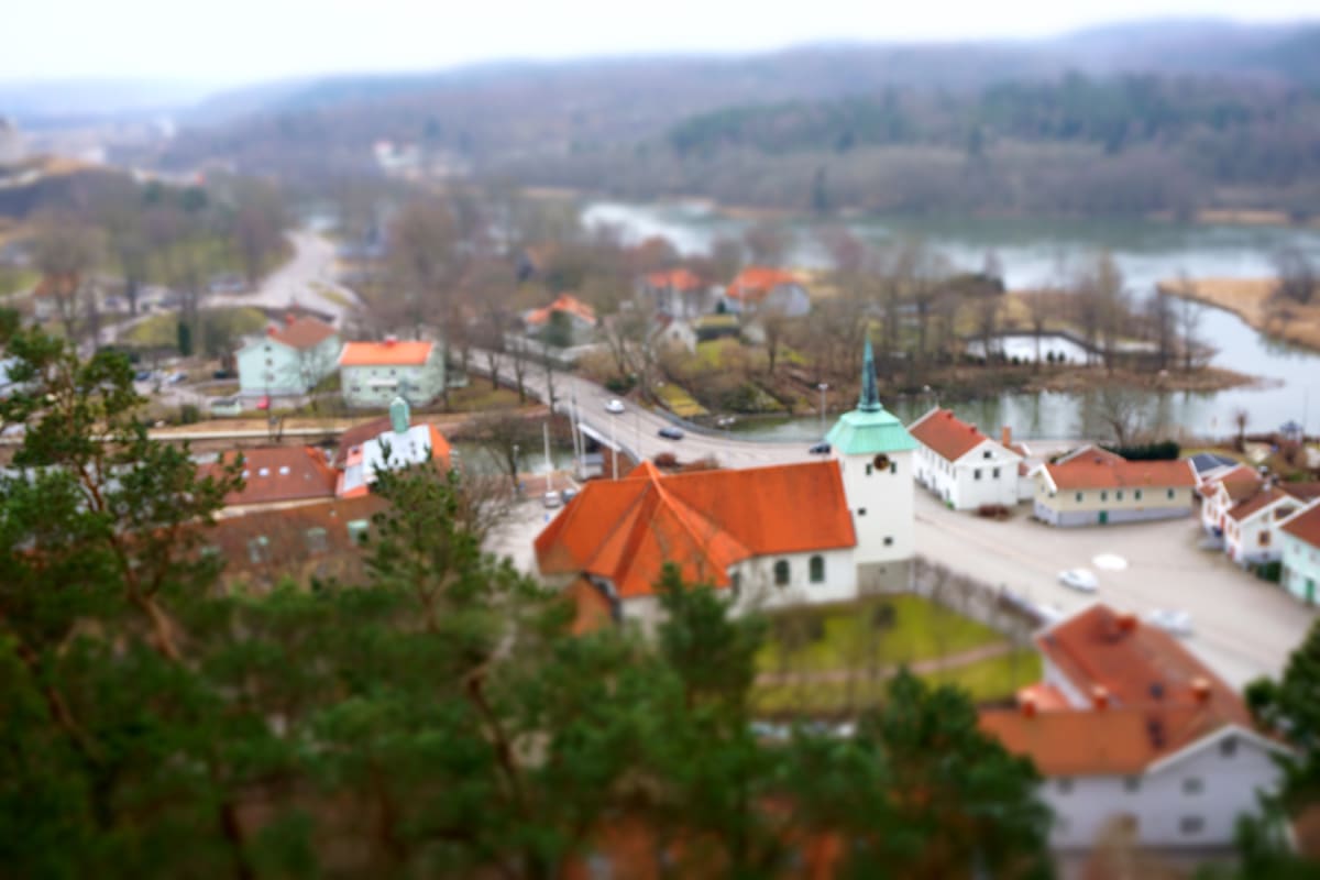 Bilden visar ett landskap med en stadskärna sedd från ovan, med en kyrka med grönt torn i förgrunden och en bro som sträcker sig över en vattenyta i bakgrunden. Omgivningen består av nakna träd och byggnader med röda tak.