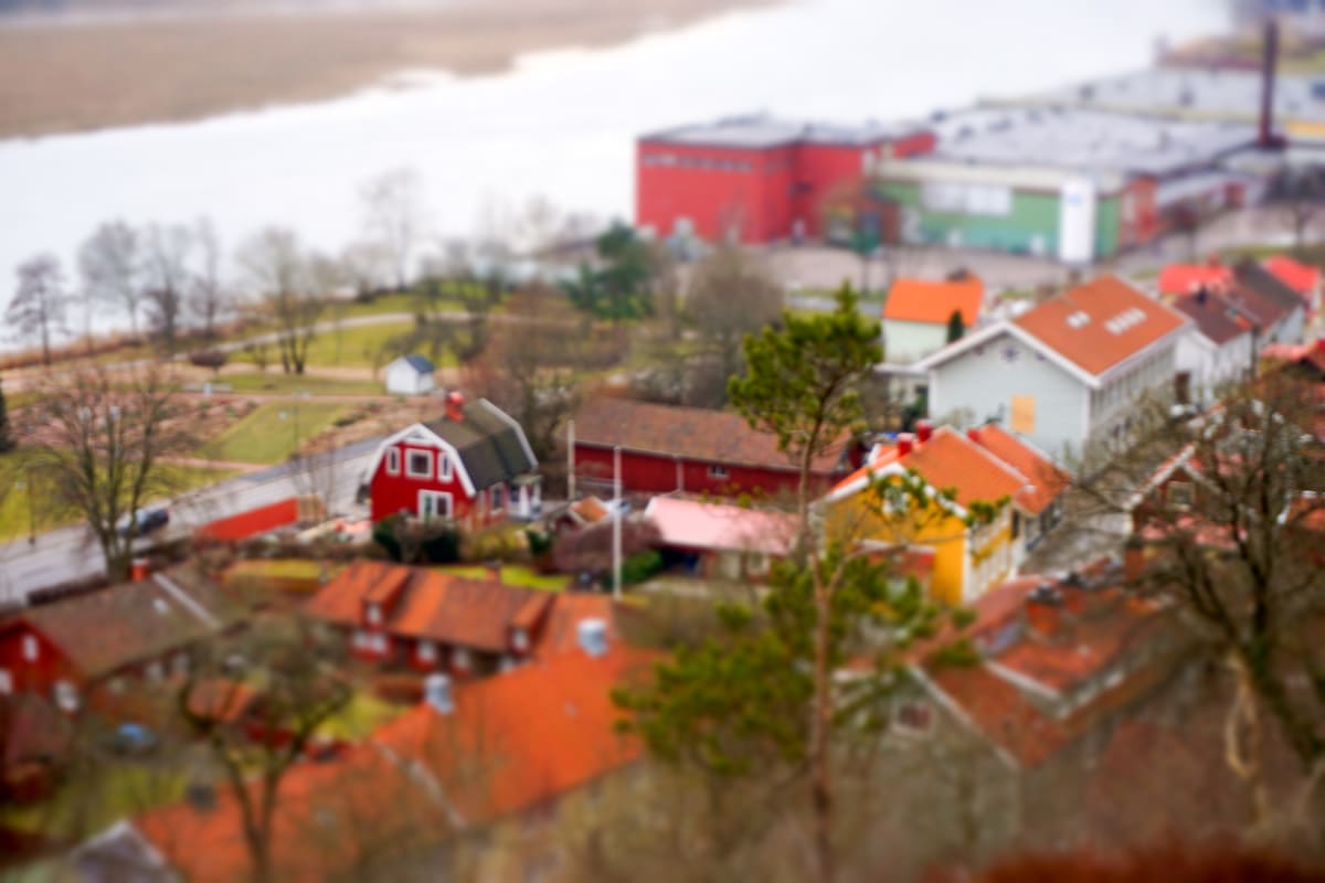 En suddig flygbild som visar ett bostadsområde med tydligt röda och orange tak. I bakgrunden syns skog, en vattenyta och industriella byggnader. Det är en ljus men molnig dag. Bilden har en tilt-shift-effekt.
