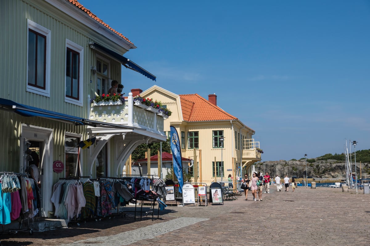 En bild som visar en livlig hamnplats med flera byggnader längs en kullerstensbelagd gata. Människor promenerar i soligt väder och kläder hänger på ställningar utanför butiker. I bakgrunden syns klippor och båtar vid vattnet.