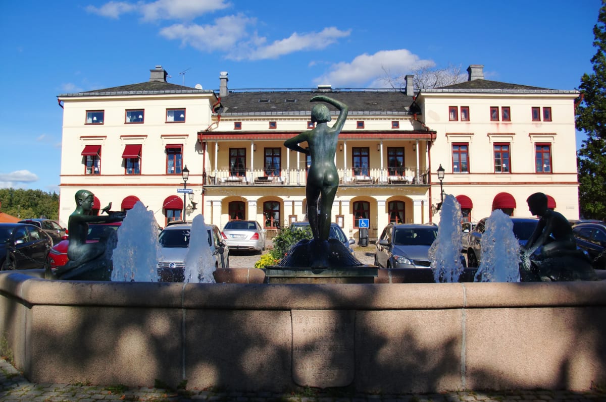 En stor, ljus byggnad med en lång balkong och röda markiser på fönstren syns i mitten av bilden. Framför byggnaden finns en fontän med statyer och vattenstrålar. Runt fontänen står parkerade bilar och himlen är blå med några vita moln.